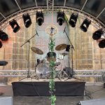 Erhöht aufgebautes Schlagzeug hinter einem mit Efeu dekorierten Mikrofonständer auf einer Veranstaltungsbühne mit halbrundem Dach. Im Hintergrund ein großes, hohes Eingangsportal in ein Sandsteingebäude.